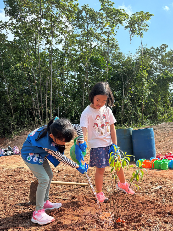 Two young girls plant a tree. One waters it from a green can, the other watches. Barrels and watering cans behind them. Trees in background.